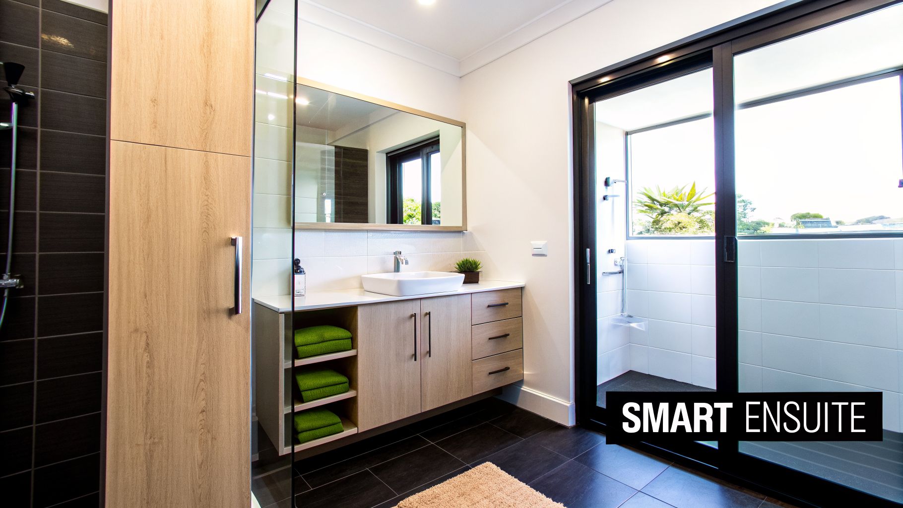 Modern ensuite bathroom with light wood vanity, vessel sink, mirror, glass shower, and green towels.